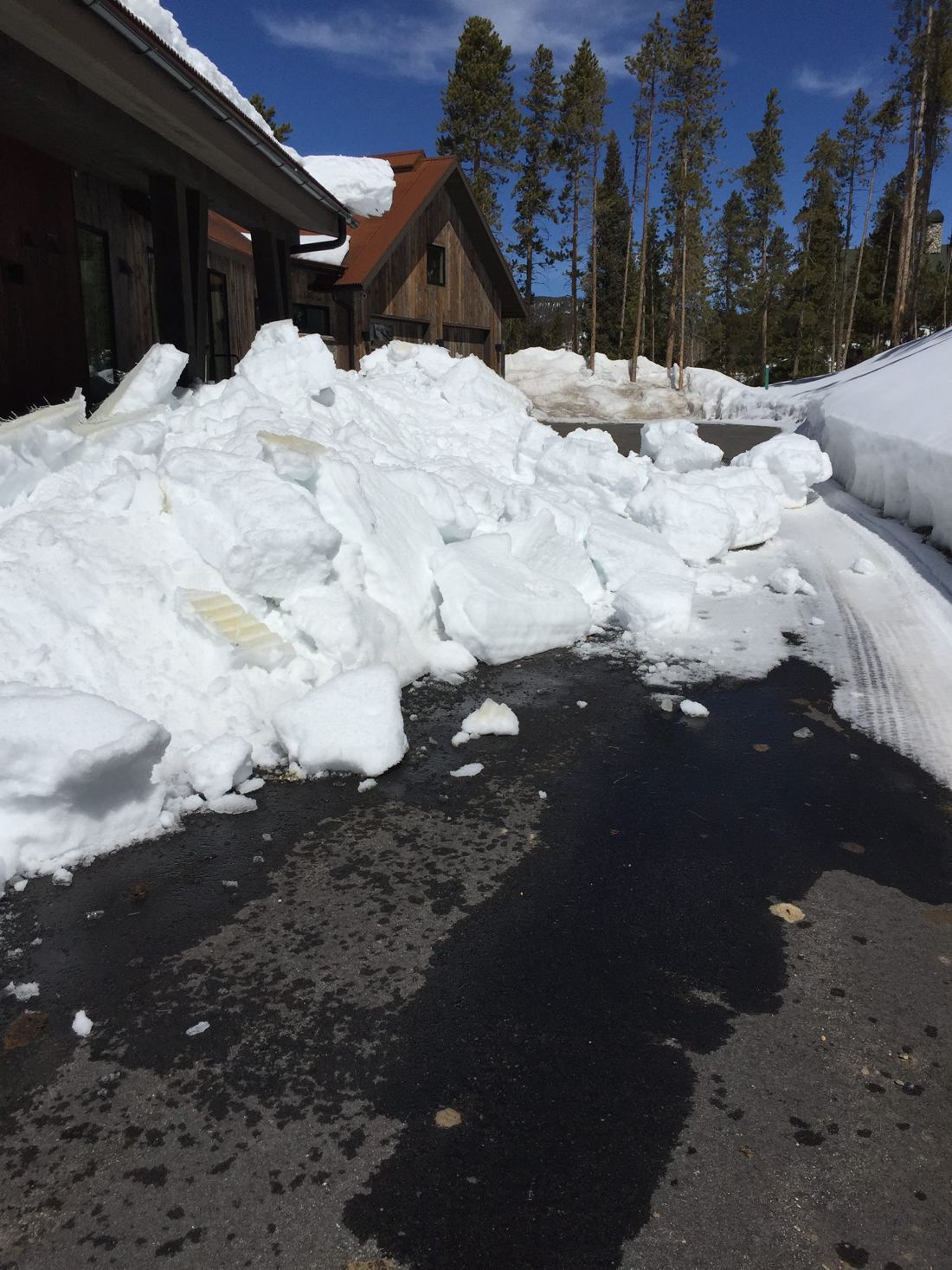 A large pile of snow and ice cleared from the asphalt driveway beside a timber and rust-roofed mountain home