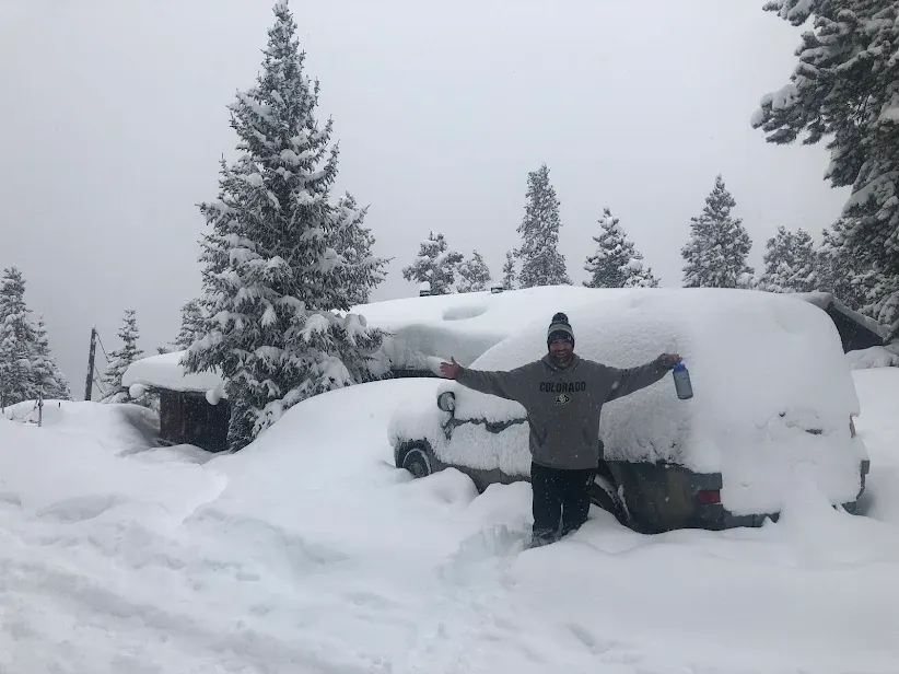 A person standing with arms out in deep snow between a snow-covered vehicle and a cabin